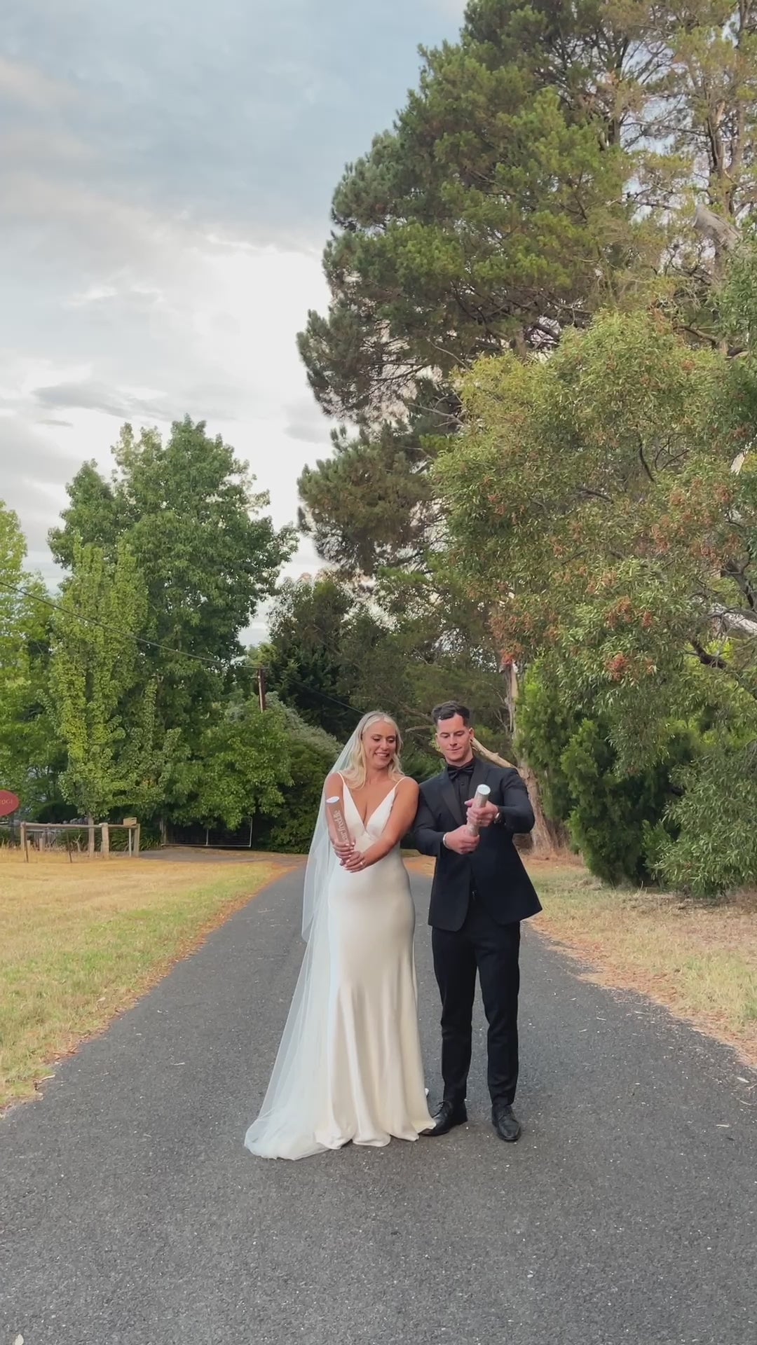 A newlywed couple laughs as they set off biodegradable confetti cannons, filling the air with pink and white confetti on a scenic road.